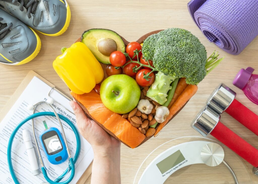 Heart shape cutting board fill with avocadi, tomato, broccoli, green apple, carrot, salmon, almonds, garlic and yellow bell pepper, sorrunded with training shoes, bottle of watter, yoga mat, a glucometer and a scale