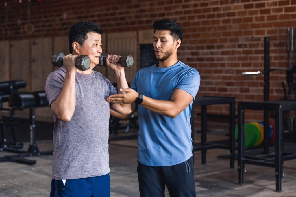 A mid adult male personal trainer helps an active senior man use hand weights 