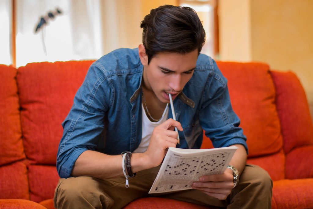 Young man sitting doing a crossword puzzle looking thoughtfully 