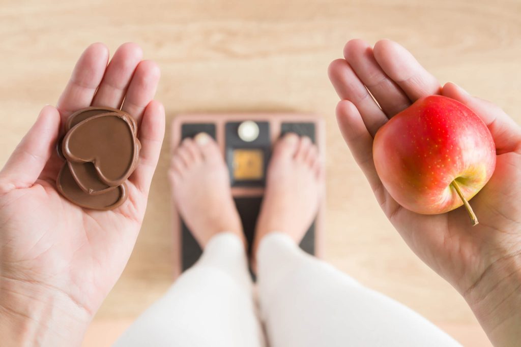 Woman standing on scales and holding apple and chocolate hearts. New start for healthy nutrition, body slimming, weight loss