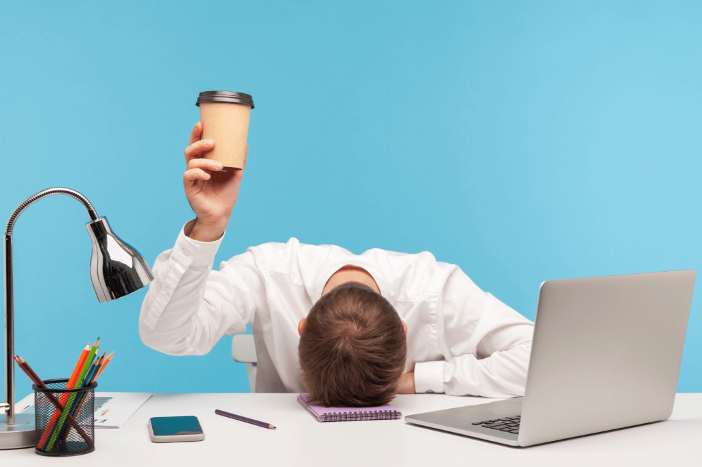 men with his head on his work desk rising a cup of coffee 