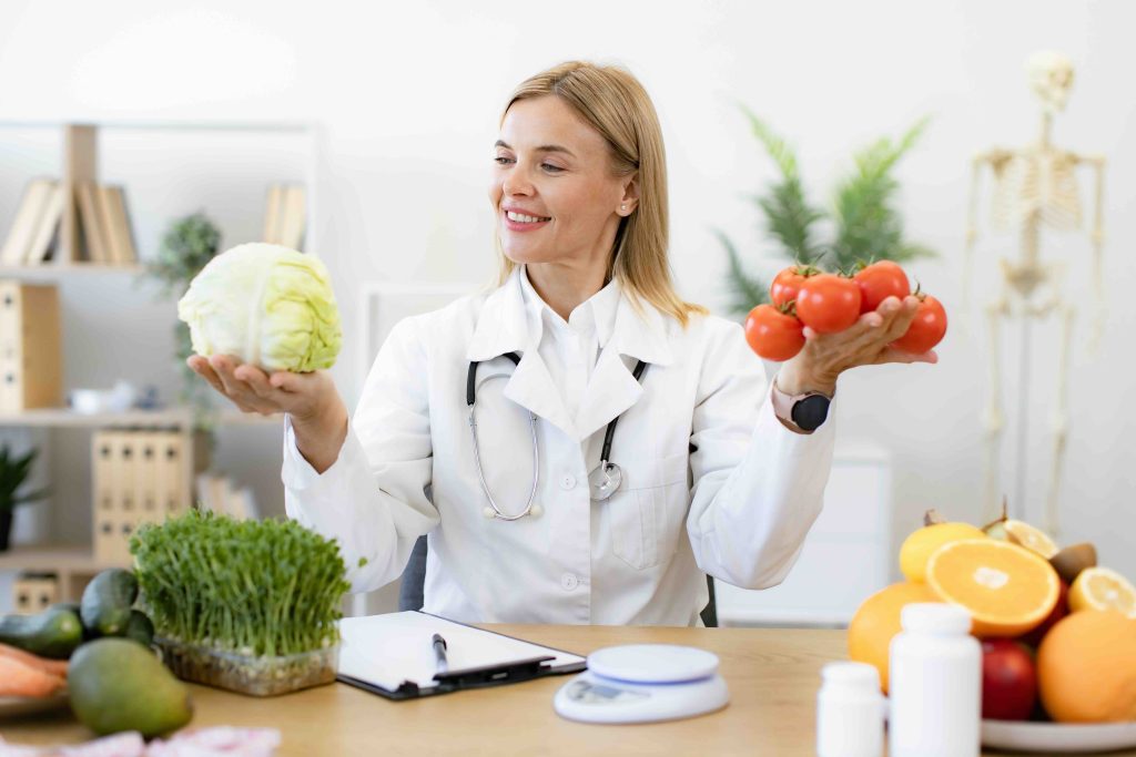 Mature female doctor nutritionist holds fresh organic tomatoes and cabbage in hands while sitting at table in office.