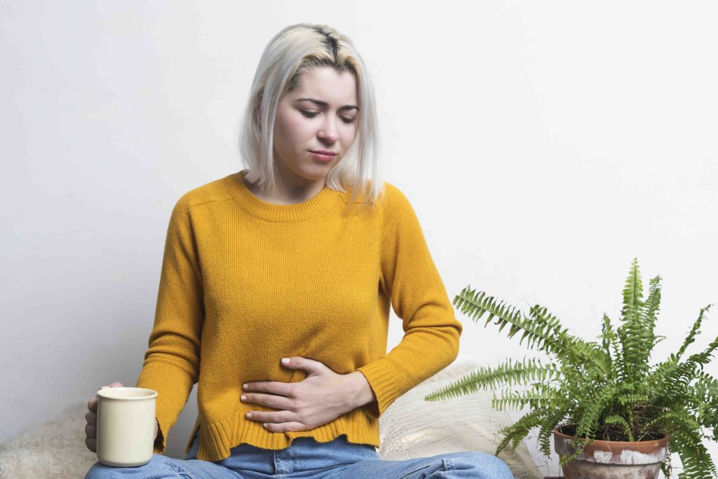 Blonde woman with nausea sitting on ground holding cup of tea and one hand on belly