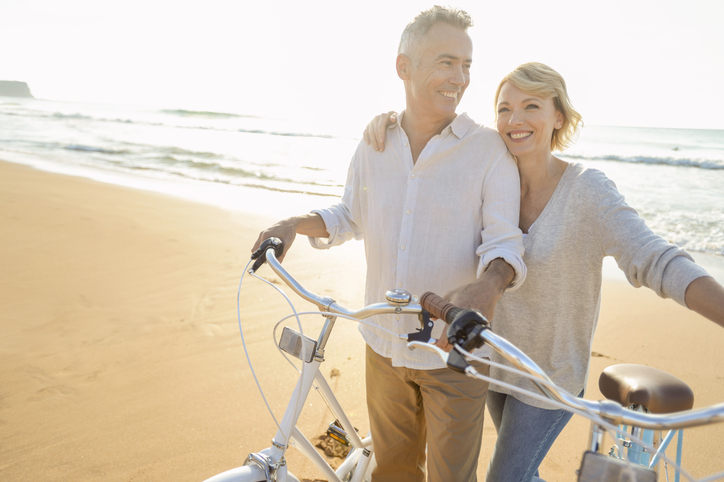 Mature couple cycling on the beach at sunset or sunrise. The ocean is in the background. They are happy and smiling. They are standing beside their bicycles. They are casually dressed.