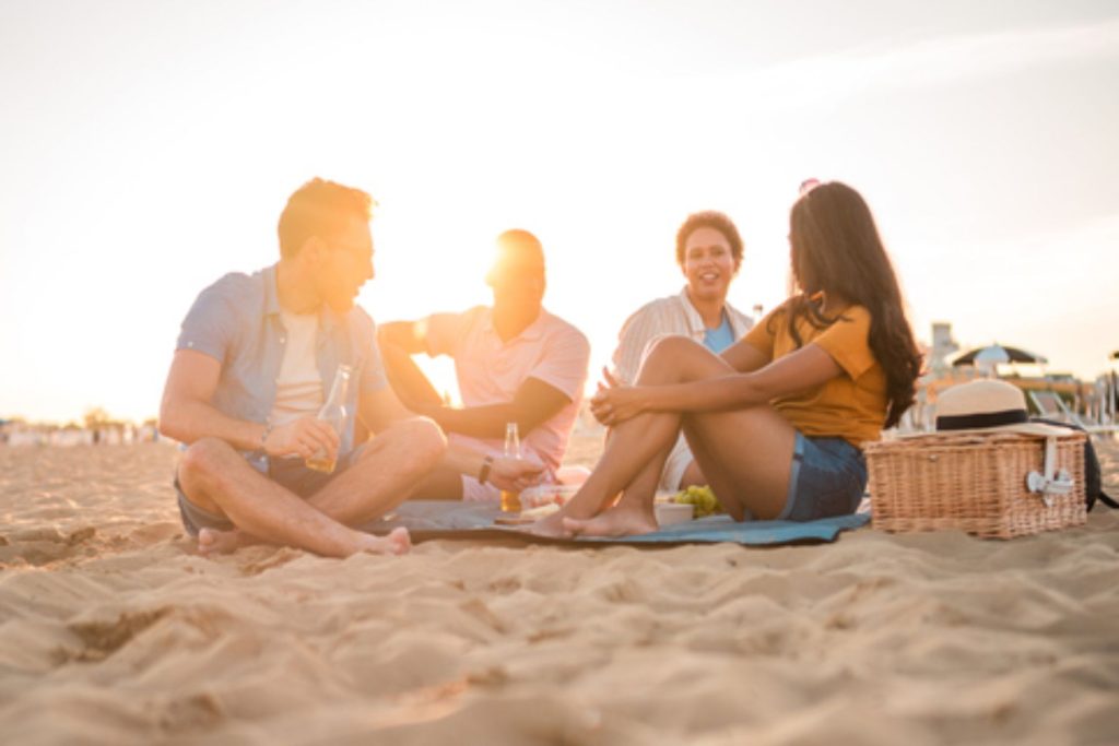 A relaxing sunset picnic on the beach with two multiracial couples. They chat, build bonds, and enjoy beer and snacks.