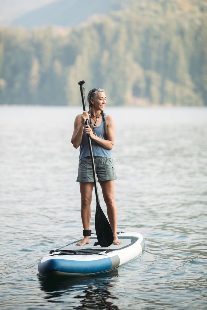 Woman with gray and brown hair in braids smiling while standing on a paddleboard on a lake