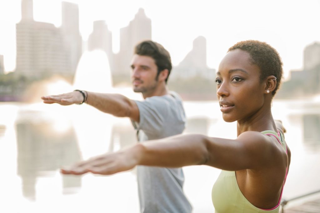 Black woman with short hair and white man in warrior two yoga pose with river and city in the background