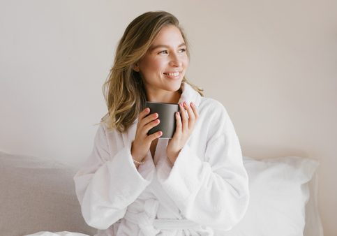 Young beautiful woman wearing white bathrobe having breakfast in bed with coffee and croissant and fresh fruits in cozy bedroom. Morning rituals.