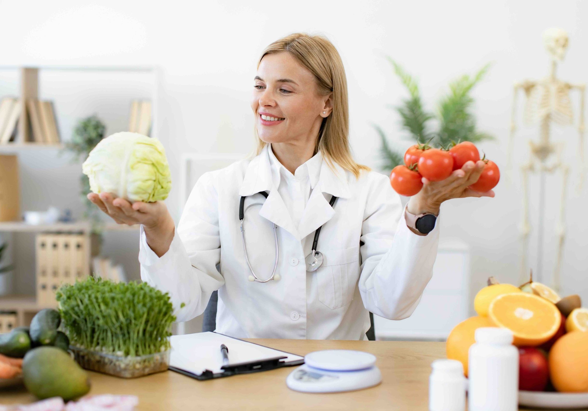 Mature female doctor nutritionist holds fresh organic tomatoes and cabbage in hands while sitting at table in office. Caucasian woman makes diet of healthy food for treatment of gastritis.