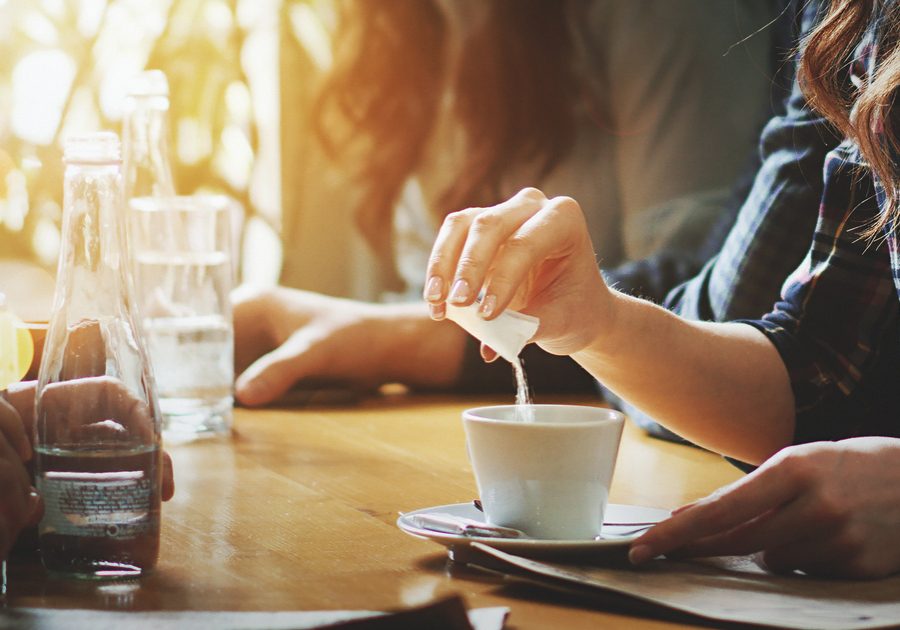 Closeup of unrecognizable woman putting some sugar into her coffee. She's having a tea spoon of sugar packed in a little bag. Blurry people in background.