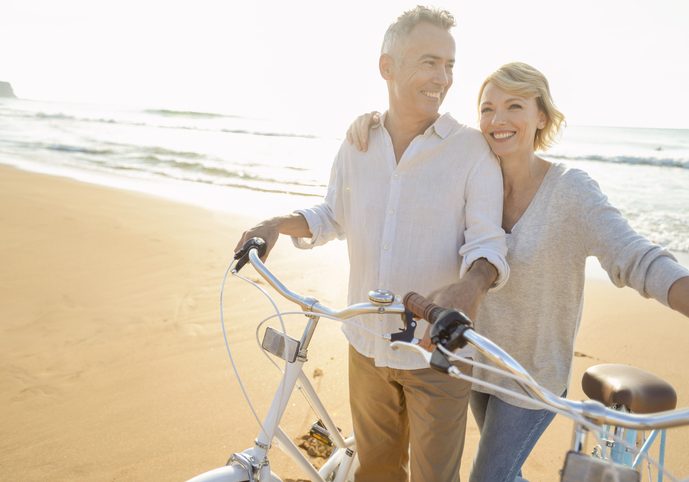 Mature couple cycling on the beach at sunset or sunrise. The ocean is in the background. They are happy and smiling. They are standing beside their bicycles. They are casually dressed. Could be a romantic retirement vacation.