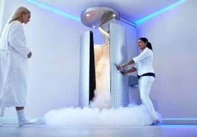 Full length portrait of nurse opening the door of cryosauna, with woman in bathrobe at the clinic. Female going for cryotherapy treatment in freezing cabinet.